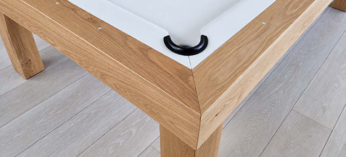 Close-up of the Rift pool table’s mitered white oak corner, black pocket, white felt, and rail sights.